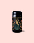 A black tea canister labeled “Magic Hour,” adorned with an illustration of a woman surrounded by floral designs, contains Persephone’s Potion: Pomegranate & Jasmine Green Tea. Infused with delicate notes of jasmine green tea and pomegranate, it offers a truly enchanting experience. The background is light pink.