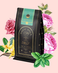 A black and green Mantra Mint™ Herbal Tea package labeled "Magic Hour" is surrounded by illustrated pink roses, yellow vanilla flowers, and green mint leaves on a pale pink background. This herbal blend is caffeine-free.