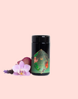 A black jar with a colorful geometric label sits beside a pink orchid, red petal, and lavender on light pink—ideal for storing Moonlight in Marrakesh: Spiced Mint Green Tea.
