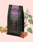 A black and gold bag of Lucid Dreams: Tulsi-Turmeric Herbal Tea for Sleep & Calm is shown with turmeric, cinnamon sticks, and herbs—ideal for those wanting a soothing tulsi tea or caffeine-free chai—on a light pink background.
