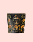 A decorative tea package features an illustrated woman in a dress among sunflowers with floral and celestial designs on black, set against soft pink. The label reads "The Sun: Energizing Adaptogenic Herbal Tea with Yaupon, Moringa & Turmeric.