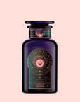 A dark purple glass jar with a label showing an eye, radiant lines, and two hands. The label reads "Queen of Wellness: Women's Hormone Balancing Tea for PMS, Healthy Cycles & Menopause." Set on a light pink background.