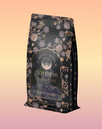 A decorative black bag of Goddess of Earl: Lady Luck Oriental Beauty Oolong Tea, featuring elegant gold and lavender floral designs, is displayed against a soft pastel gradient background.