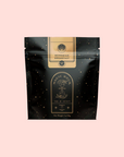 A black resealable pouch labeled "Magic Hour" and "Bohemian Breakfast Vanilla Puerh Black Tea for Digestion & Energy," with mystical designs and gold accents, stands against a pale pink background. This organic blend is a luxurious coffee alternative.
