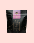 A black resealable pouch with gold speckles labeled "Child’s Pose™ Herbal Adaptogen Tea Blend for Sleep & Restful Calm," featuring an illustrated hourglass and cup with light purple and gold accents, set on a pale pink background.