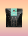 A black, starry tea bag labeled "Mantra Mint™ Herbal Tea" stands before a pastel pink-orange gradient. Its celestial packaging has a mint-green label and notes that this blend is caffeine-free.