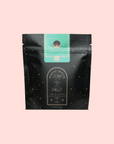 A black resealable pouch with gold stars and a green label displays “Mantra Mint™ Herbal Tea.” Below, a circular emblem reads “Magic Hour: Time ∞ Infinity.” The caffeine-free tea sits against a light pastel pink backdrop.