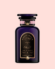 A sleek dark purple glass bottle with a black cap holds "Almond Matcha Green Tea for Joy." The black and gold label features pink and gold accents, set against a pale pink background.
