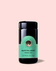 A black jar of Mantra Mint™ Herbal Tea, a caffeine-free blend with Ashwagandha and a green label with a circular logo, is centered on a light pink background.