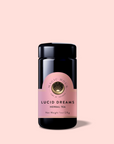 A dark glass jar with a pink label reading "Lucid Dreams: Tulsi-Turmeric Herbal Tea for Sleep & Calm" and a gold emblem sits upright and centered against a light pink background, perfect for relaxing like a caffeine-free chai or calming tulsi tea.