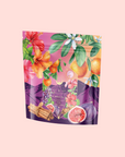 The Pink Moment Pixie - Hibiscus Tangerine Dream Herbal Tea comes in a vibrant package with floral and fruit designs featuring elderberry, figs, grapes, cinnamon sticks, and green leaves on a pink background. Naturally caffeine free.