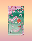 The Queen of Kyoto Sencha Green Tea package features pink Japanese Cherry Blossoms, white cranes, and a hexagonal label on green, set against a peach-to-yellow gradient background.