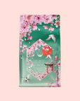 A green and pink tea bag of Queen of Kyoto features cherry blossoms, flying cranes, and floral art inspired by the Japanese Cherry Blossom Festival with "Green Tea, Queen of Kyoto" text on a soft pink background.