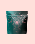 A dark green resealable pouch with pink mystical line art shows the label “Queen of Wellness: Women's Hormone Balancing Tea for PMS, Healthy Cycles & Menopause,” promoting women’s wellness, against a pale pink background.
