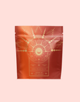 A red-orange tea package with gold celestial motifs—rays, stars, and hands—is labeled “Queen of the Harvest: Herbal Relaxation Tea.” This organic herbal blend stands out against a solid light pink background.