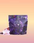 A purple pouch of Amethyst: Strawberry-Passionfruit Gemstone Wellness Tea, with an amethyst gemstone design, stands on a peach-yellow gradient beside a pink orchid and lavender—ideal for those seeking spiritual clarity.