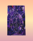A purple tea package labeled "Amethyst: Strawberry-Passionfruit Gemstone Wellness Tea" stands upright against a soft orange-pink gradient. Its geometric design features a large faceted gem, emphasizing the herbal tea's spiritual wellness focus.