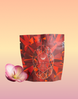 A package of Carnelian: Caffeine-Free Apricot Amaretto Tea with a red geometric design stands upright on a pastel gradient background, beside a single pink orchid blossom.