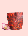 A red and orange geometric-patterned Carnelian: Caffeine-Free Apricot Amaretto Tea package stands upright beside a single pink flower on a pale pink background.