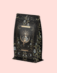 A decorative black tea bag with gold and blue floral designs and a mystical figure, labeled "The High Priestess Ginger-Mint-Cardamom Tea—Powerful Dreams & Illuminated Insights" on a pale pink background.