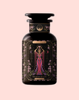 A dark glass bottle labeled "The Queen of Cups: Hibiscus-Vanilla-Berry Tea for Balance & Heart Health" features ornate art of a regal woman in pink with celestial symbols, celebrating balance and heart health on a light pink background.