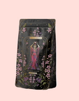 A tea package labeled “The Queen of Cups: Hibiscus-Vanilla-Berry Tea for Balance & Heart Health” features floral designs and a woman in a pink gown, set against a pale pink background, and promotes emotional balance.
