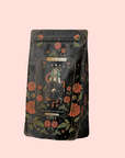 A black tea bag package with gold and red floral designs and a "The Wheel of Fortune" tarot card illustration showcases The Wheel of Fortune: Ginseng Spice Tea for Adapting to Change, an adaptogenic blend, set against a pale pink background.