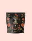 A black tea pouch with ornate floral and star designs, featuring "The Wheel of Fortune: Ginseng Spice Tea for Adapting to Change" by Magic Hour—a tarot-inspired adaptogenic wellness blend on a pale pink background.