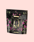 A black, resealable pouch with gold, green, and pink floral designs. The label reads "Magic Hour" and "The Lovers Hibiscus-Cinnamon-Vanilla Love Potion," with two figures among flowers and stars—infused with Damiana. Pale pink background.