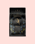 A black coffee bag labeled "Aquarius: Visionary Goddess Tea" features a celestial gold zodiac figure, sun rays, blue flowers, and stars on a light pink background—ideal for fans of Organic Golden Monkey Tip Black Tea.