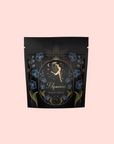 A black resealable pouch labeled "Aquarius: Visionary Goddess Tea" showcases gold and blue florals, celestial motifs, and a woman pouring water under stars—evoking the rich Yunnan tea within—set against a pale pink backdrop.