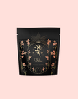 A black resealable pouch with gold and pink floral designs, featuring a gold figure holding scales, labeled “Libra Shatavari-Cardamom-Rose White Love Cake Tea,” evokes the enchanting aroma of rose and pistachio.