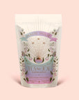 A cream-colored pouch labeled "Tea for Joy: Almond Matcha: Green Matcha Tea with Almonds" adorned with floral and celestial designs rests on a pale pink background. The packaging showcases an antioxidant-rich ceremonial green matcha tea blend featuring almonds and love.