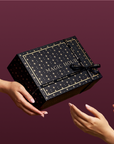 A person hands a Branded Gift Box—black and adorned with gold stars and the words “MAGIC HOUR”—to another person, creating a memorable gifting moment against a rich burgundy background.
