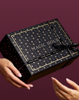 A person hands a Branded Gift Box—black and adorned with gold stars and the words “MAGIC HOUR”—to another person, creating a memorable gifting moment against a rich burgundy background.