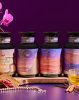 The Balancing Dosha Bundle—four jars of caffeine-free Ayurvedic tea with unique labels—rests on a wooden tray surrounded by an orchid, tea accessories, jewelry, and a glass of tea against a purple backdrop.