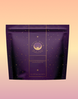 A purple Perfumes of Provence: Lavender Bouquet Tea pouch for calm moods and beautiful skin, with gold stars and a circular moon emblem on elegant packaging, set against a soft peach-pink gradient background.