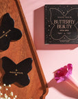 A flatlay showcasing butterfly-shaped black jade Butterfly Beauty Gua Sha tools by Magic Hour, one on a wooden tray and one beside a boxed set labeled "Magic Hour Butterfly Beauty." A single pink calla lily and white flowers accent the pink background, evoking the serene elegance of Magic Hour Tea.