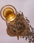 A glass cup filled with a light amber liquid sits on a light pink surface next to a golden leaf-shaped tray. Loose Magic Hour Gunpowder Green Tea leaves are scattered from the tray onto the surface, adding an earthy flavor to the scene. The arrangement creates an aesthetically pleasing and artistic presentation.
