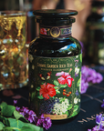 A decorative bottle of Magic Hour Hibiscus Elderberry: Cosmic Garden Iced Tea sits on a table surrounded by purple flowers. The label, showcasing vibrant illustrations of hibiscus flowers, elderberries, butterflies, and foliage, gives it a magical and whimsical appearance reminiscent of Organic Tea at Magic Hour.