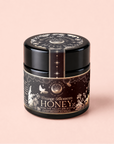A black glass jar of Orange Blossom Honey: Raw Ojai Honey from the Valley of the Moon, made with organic California honey, featuring floral and bird art on its label, set on a soft pink background.