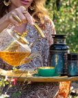 A woman pours Hathor of Joy: Honey Nectar Herbal Tea from a glass teapot into a green teacup on a tray, with tea containers beside her. She wears patterned clothes and gold jewelry, sitting outdoors among flowers and enveloped in the gentle fragrance of honey nectar.