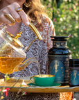 A woman pours Hathor of Joy: Honey Nectar Herbal Tea from a glass teapot into a green teacup on a tray, with tea containers beside her. She wears patterned clothes and gold jewelry, sitting outdoors among flowers and enveloped in the gentle fragrance of honey nectar.