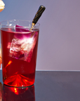 A glass of deep red cocktail featuring a large ice cube rests on a sleek surface. A black stirrer blends in, infusing delicate herbal notes from the Heart Health Tea Sampler Set. In the background, gentle pink petals and a gradient purple wall contribute elegance and an aura of cardiovascular wellness.