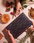 Hands holding an Alchemy of Autumn Box, a black rectangular tea box labeled “Magic Hour” in gold, surrounded by a glass teapot, a gold teapot, berries, and a small fruit tart.