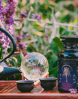 A hand pours tea from a black teapot into cups beside a dark floral jar labeled “Monthly Magic Tea Journey Subscription,” evoking ritual and Divine Feminine exploration amid purple flowers and lush greenery outdoors.