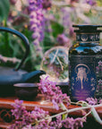 A decorative tea bottle from the Monthly Magic Tea Journey Subscription sits among purple flowers, a teapot, and a teacup outdoors—ideal for goddess ritual tea beneath lush greenery and sunlight.