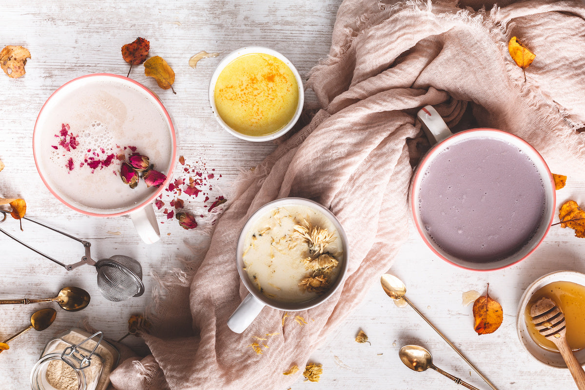 An assortment of four colorful lattes in mugs on a white wooden surface. A pink scarf weaves among the mugs, surrounded by spoons, a tea infuser, scattered dried flowers, and leaves. The lattes are topped with various decorative ingredients.