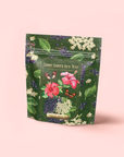 A green floral package of "Hibiscus Elderberry: Cosmic Garden Iced Tea" displays vibrant hibiscus flowers, elderberries, and leaves on a pink background, highlighting this antioxidant-rich iced tea blend.