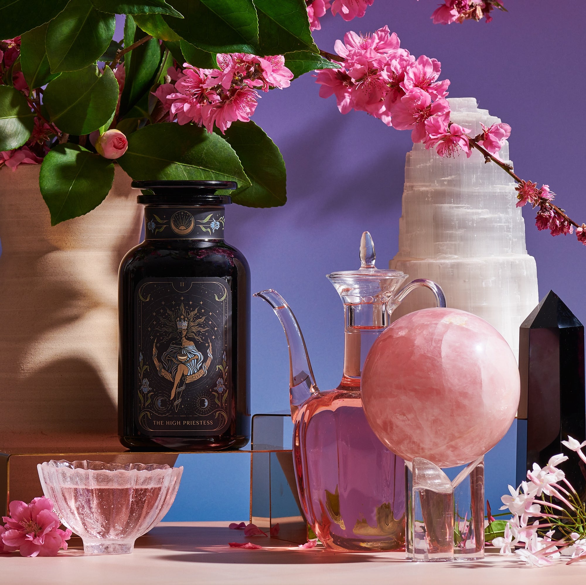 The Tea and Tarot to Go Gift Set from Magic Hour presents a purple-themed arrangement that includes a dark glass bottle, a clear glass pitcher filled with pink liquid, a rose quartz sphere, and various crystal formations surrounded by pink cherry blossoms, all set against a purple background to evoke the Tarot archetype.